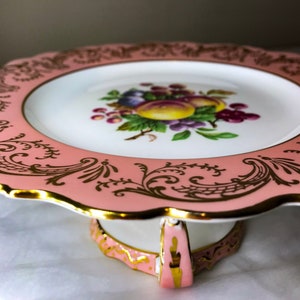 Puede incluir: Un soporte para pasteles de porcelana rosa y dorado con borde festoneado. El plato presenta un diseño de frutas en el centro y filigranas doradas alrededor del borde. El soporte tiene una base decorativa con detalles dorados.