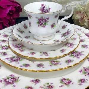 Puede incluir: Un juego de cuatro platos de porcelana blanca con un delicado patrón floral con violetas moradas y un borde dorado. El juego incluye una taza de té y un platillo.