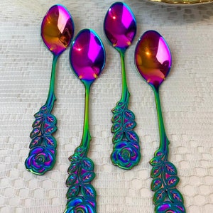 Puede incluir: Cuatro cucharas iridiscentes con detalles de rosas y hojas en los mangos. Las cucharas tienen un vibrante degradado de color púrpura, rosa y verde. Una taza y un platillo florales están en el fondo.