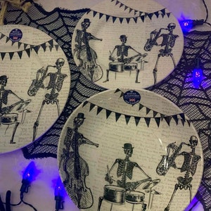 May include: Three white ceramic dinner plates with black skeleton band illustrations. The plates feature a black and white patterned background with a banner that reads "Royal Stafford".