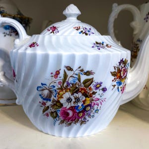 Może przedstawiać: Biały porcelanowy imbryk z spiralnym wzorem i kwiatowymi akcentami. Imbryk posiada zakrzywioną wylewkę, pętlę uchwytu i pokrywkę z guzikiem. Kwiatowy wzór zawiera różowe, niebieskie i żółte kwiaty.