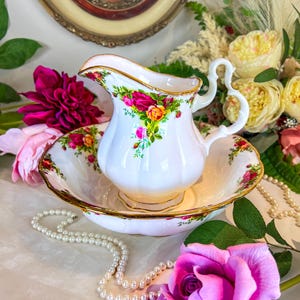 May include: A white porcelain pitcher and bowl set with a floral rose pattern. The set has gold trim and is surrounded by flowers and a pearl necklace. The pitcher is approximately 6 inches tall.