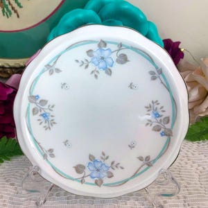 May include: White ceramic plate with a scalloped edge, featuring a light blue floral design and a teal border. The plate has a gold trim and is displayed on a clear stand. The floral pattern includes blue flowers and gray leaves.