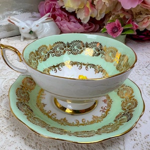 May include: A vintage teacup and saucer set. The teacup and saucer are light green with gold filigree and trim. The teacup has a white interior with a yellow floral design. The handle and rim are gold.