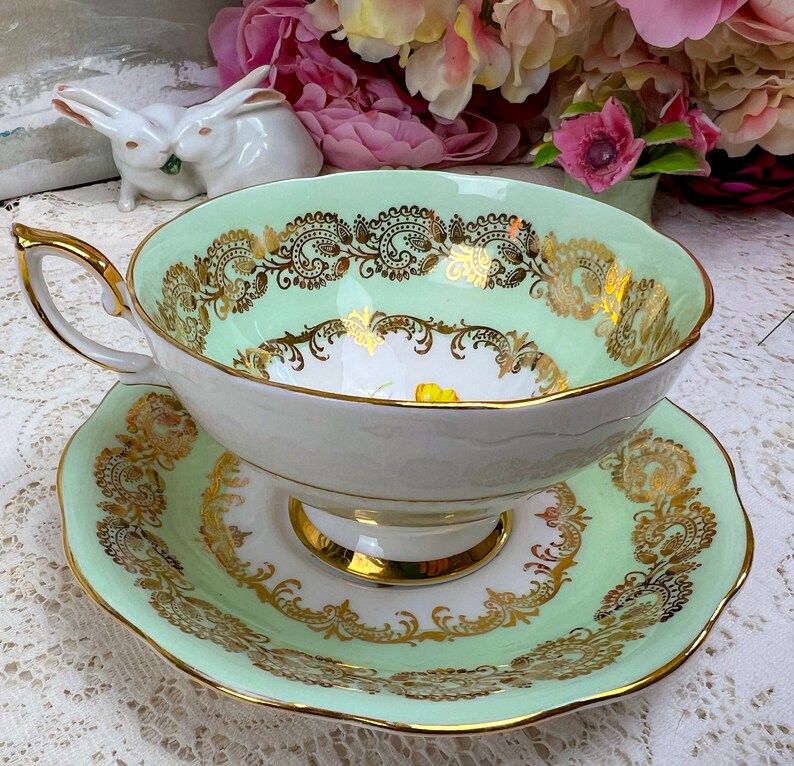 May include: A vintage teacup and saucer set. The teacup and saucer are light green with gold filigree and trim. The teacup has a white interior with a yellow floral design. The handle and rim are gold.