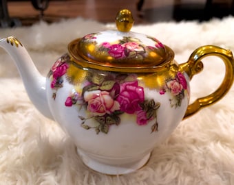 Może przedstawiać: Biały porcelanowy czajnik z pozłacanym wykończeniem i kwiatowym wzorem z różowymi różami. Czajnik ma pokrywkę z pozłacanym uchwytem.