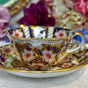 Puede incluir: Un juego de taza y platillo detallado. La porcelana blanca está decorada con un estampado floral en rojo, naranja y dorado, con detalles azules y dorados. El borde y el asa están dorados. Un juego de té clásico.