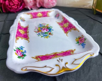Puede incluir: Una bandeja de porcelana blanca con un diseño floral rosa y dorado. La bandeja tiene un borde festoneado y un ribete dorado.