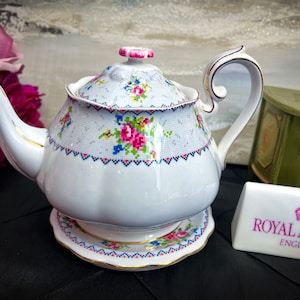 May include: A white teapot with a floral pattern and a matching saucer. The teapot has a lid with a pink knob and a gold trim. The saucer has a gold rim and a floral pattern. The teapot is sitting on a black surface. The text "ROYAL ALBERT ENGLAND" is visible on a white sign.