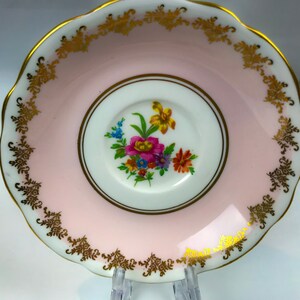 Puede incluir: Un platillo rosa con un borde dorado y un dise&ntilde;o floral en el centro. El platillo est&aacute; hecho de porcelana y tiene un borde festoneado.