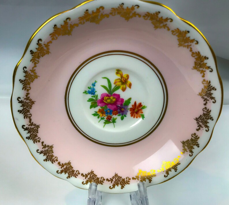 Puede incluir: Un platillo rosa con un borde dorado y un dise&ntilde;o floral en el centro. El platillo est&aacute; hecho de porcelana y tiene un borde festoneado.