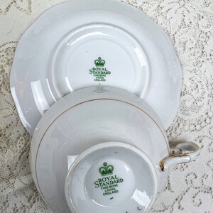 May include: White Royal Standard fine bone china teacup and saucer set. The teacup and saucer have a gold trim. The bottom of the teacup and saucer have the Royal Standard logo in green. The teacup has a handle.