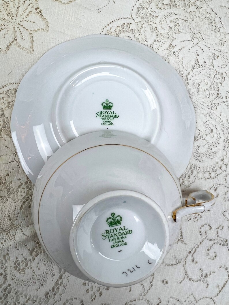 May include: White Royal Standard fine bone china teacup and saucer set. The teacup and saucer have a gold trim. The bottom of the teacup and saucer have the Royal Standard logo in green. The teacup has a handle.