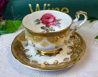 May include: A vintage teacup and saucer set. The white porcelain cup features a vibrant pink rose and gold filigree detailing. The saucer mirrors the cup's design with a gold rim and matching floral accents. A classic tea set.