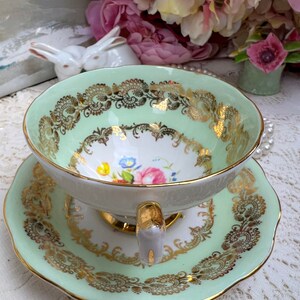 May include: A vintage teacup and saucer set. The cup and saucer are a soft green with gold filigree and trim. The interior of the cup features a floral design with pink, blue, and yellow flowers. The handle is gold.