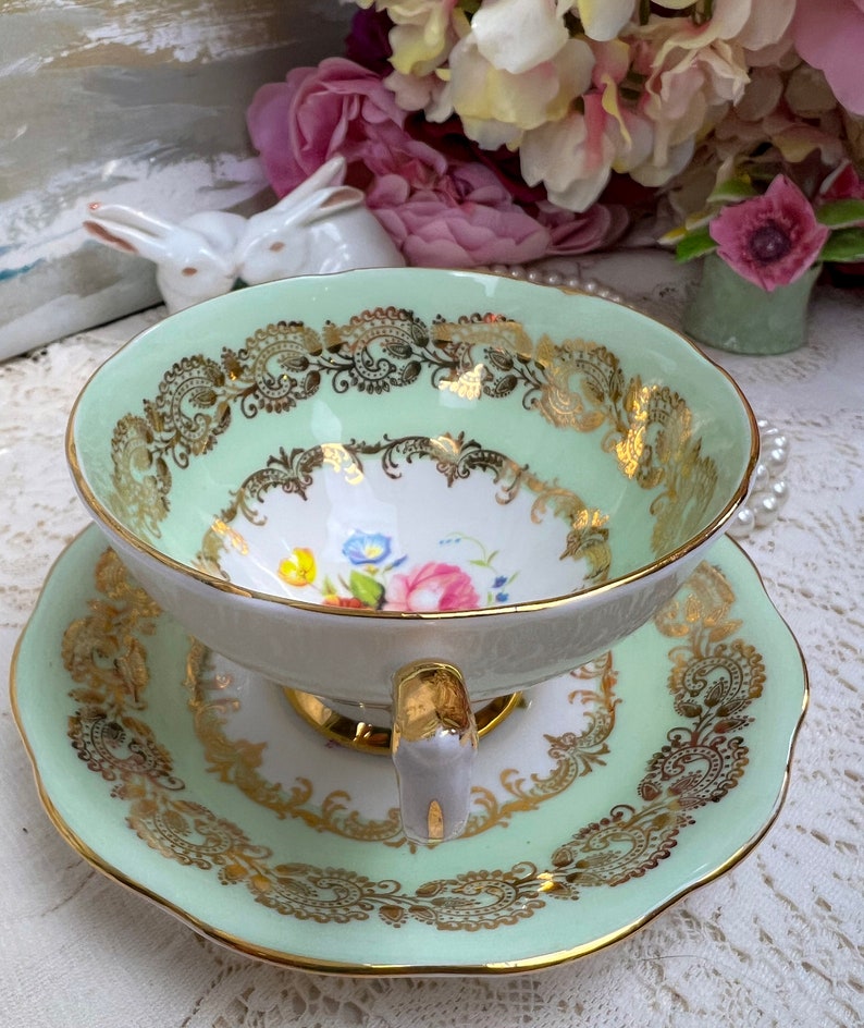 May include: A vintage teacup and saucer set. The cup and saucer are a soft green with gold filigree and trim. The interior of the cup features a floral design with pink, blue, and yellow flowers. The handle is gold.