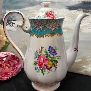 May include: A white porcelain teapot featuring a floral bouquet with pink, blue, and yellow flowers on the body. The upper rim has a teal band with white floral patterns and gold trim. It includes a matching lid with a pink rose motif.