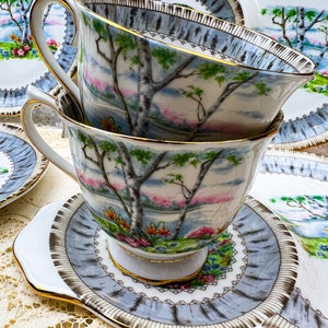 May include: Stacked teacups and saucers with a landscape design featuring trees, a lake, and colorful flowers. The teacups and saucers have a light blue and gold trim. The teacups are resting on a white and light blue saucer.