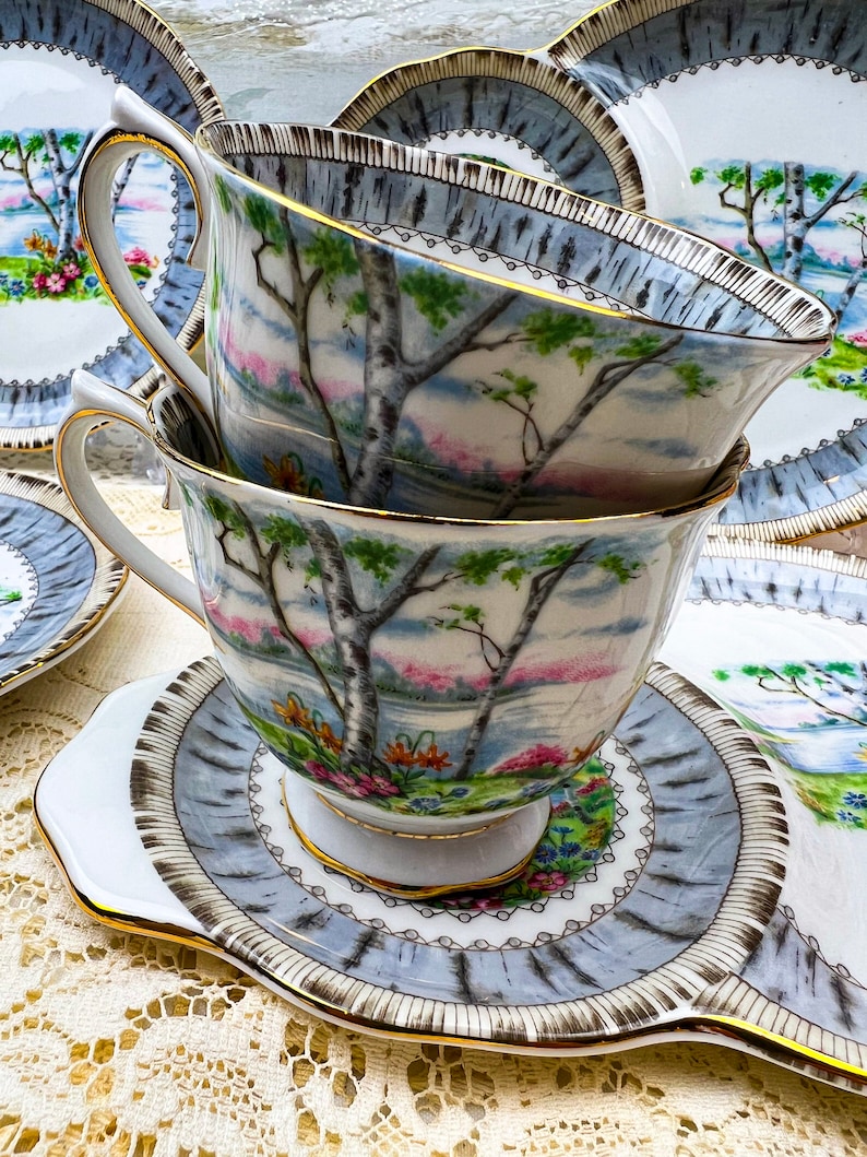 May include: Stacked teacups and saucers with a landscape design featuring trees, a lake, and colorful flowers. The teacups and saucers have a light blue and gold trim. The teacups are resting on a white and light blue saucer.