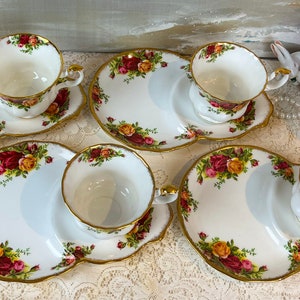 May include: Set of four white teacups and saucers with a floral pattern. Each saucer has a small plate attached for snacks. The floral pattern features red, yellow, and green roses.