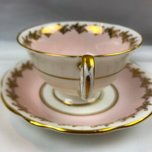 Puede incluir: Un juego de taza y platillo de t&eacute; rosa con ribetes dorados y un delicado patr&oacute;n floral. La taza tiene un interior blanco y un asa dorada.