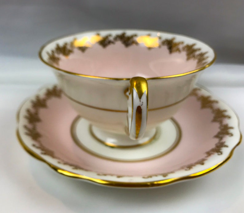 Puede incluir: Un juego de taza y platillo de t&eacute; rosa con ribetes dorados y un delicado patr&oacute;n floral. La taza tiene un interior blanco y un asa dorada.