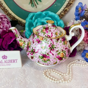 Puede incluir: Una tetera Royal Albert England con un estampado floral en rosa, amarillo y burdeos. La tetera tiene un ribete dorado y un asa blanca. Flores y un collar de perlas completan la escena.