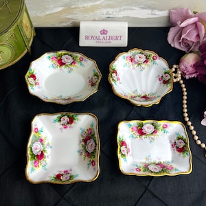 May include: Four white porcelain dishes with gold trim and floral designs. The dishes are all different shapes and sizes, and they are arranged on a black surface. The dishes are made by Royal Albert, England.