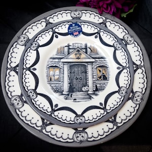 May include: A set of two gray and white ceramic dinner plates with a black and white design featuring a house with a door and windows. The plates have a scalloped edge and a black and white design of pumpkins and other Halloween motifs. The plates are made by Royal Stafford and say "1666" on the front.