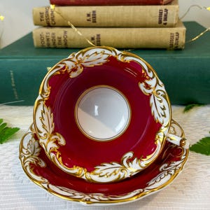 May include: A red teacup and saucer with gold trim and a white interior. The teacup and saucer feature a floral design with gold accents.
