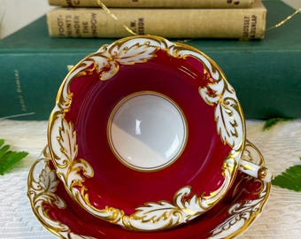 May include: A red teacup and saucer with gold trim and a white interior. The teacup and saucer have a floral design with gold accents.