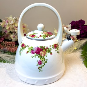 May include: White enamel teapot with a floral design featuring pink and yellow roses. The teapot has a silver lid and a white handle.