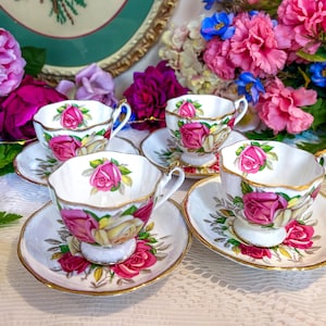 Puede incluir: Cuatro tazas y platillos blancos con un diseño de rosas. Las tazas y los platillos tienen ribetes dorados. El fondo incluye flores rosas y moradas y un plato decorativo.