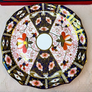 Puede incluir: Un plato de porcelana decorativo con un patrón floral azul, dorado y rojo. El plato tiene un borde festoneado y un círculo central blanco.