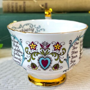 May include: A white teacup with a gold rim and floral design. The cup has a blue and green floral pattern with a red heart in the center. The text "Anniversary" is written on the rim of the cup. The text "Coral, Ruby, Sapphire, Golden, Emerald, Diamond" is written on the left side of the cup. The text "One, Two, Three, Four, Five" is written on the right side of the cup.