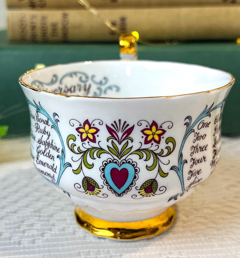 May include: A white teacup with a gold rim and floral design. The cup has a blue and green floral pattern with a red heart in the center. The text "Anniversary" is written on the rim of the cup. The text "Coral, Ruby, Sapphire, Golden, Emerald, Diamond" is written on the left side of the cup. The text "One, Two, Three, Four, Five" is written on the right side of the cup.