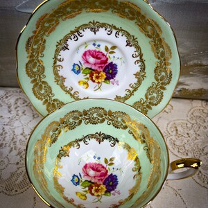May include: A vintage teacup and saucer set. The cup and saucer have a light green border with gold filigree detailing. The center of the cup and saucer features a floral design with pink, purple, yellow, and blue flowers. The handle of the cup is gold.