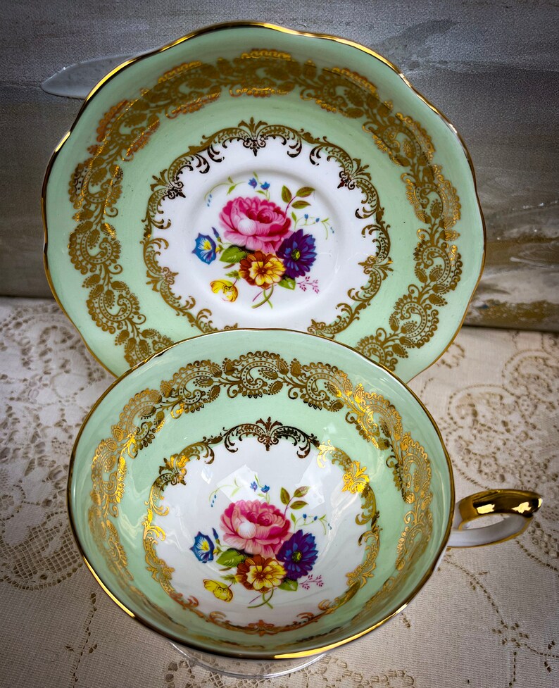 May include: A vintage teacup and saucer set. The cup and saucer have a light green border with gold filigree detailing. The center of the cup and saucer features a floral design with pink, purple, yellow, and blue flowers. The handle of the cup is gold.