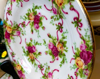 Könnte beinhalten: Ein weißer Porzellanteller mit goldfarbenem Rand zeigt ein Blumenmuster aus rosa und gelben Rosen mit grünen Blättern und rosa Bändern.