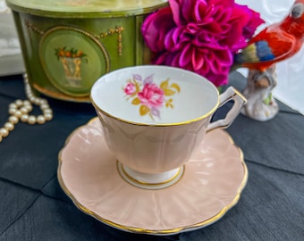 Puede incluir: Una taza de té y un platillo rosas con un diseño floral. La taza de té tiene un borde dorado y el platillo tiene un borde dorado y un borde festoneado.