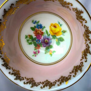 Puede incluir: Un platillo de taza de t&eacute; rosa con un borde dorado y un dise&ntilde;o floral en el centro. Las flores son amarillas, moradas, azules y rosas.