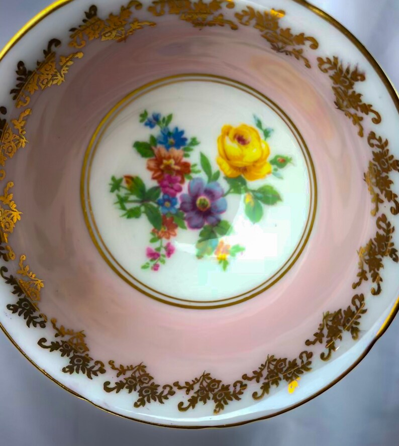 Puede incluir: Un platillo de taza de t&eacute; rosa con un borde dorado y un dise&ntilde;o floral en el centro. Las flores son amarillas, moradas, azules y rosas.