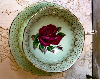 May include: A green and gold teacup and saucer set with a delicate floral design featuring a single red rose. The saucer has a scalloped edge and a repeating pattern of gold dots and diamonds.