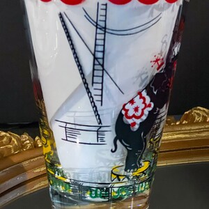 May include: A vintage glass drinking glass with a circus theme. The glass features a black and white illustration of an elephant standing on a pedestal with a red and white striped saddle. The glass has a gold rim and a white background.