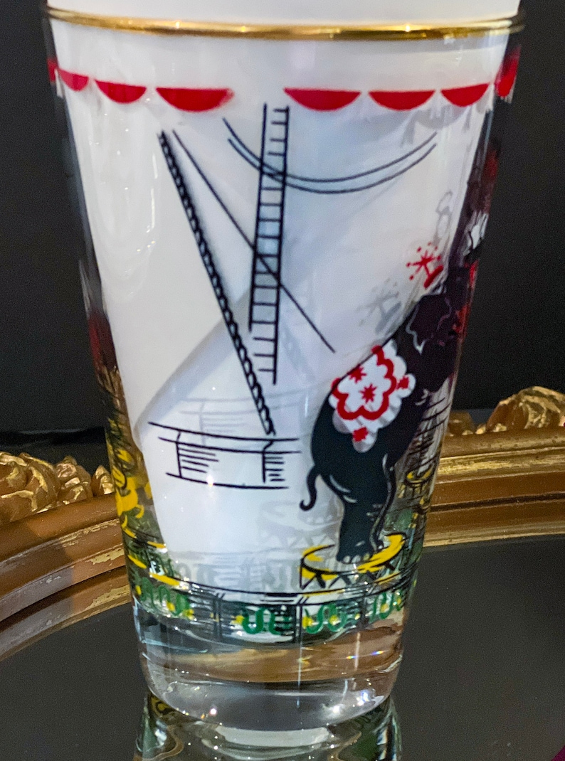 May include: A vintage glass drinking glass with a circus theme. The glass features a black and white illustration of an elephant standing on a pedestal with a red and white striped saddle. The glass has a gold rim and a white background.