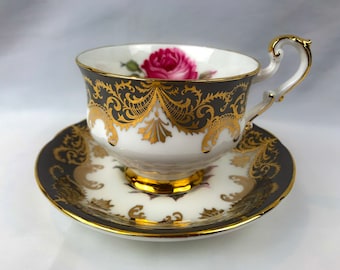 May include: A white teacup and saucer with a black and gold floral design. The cup has a pink rose painted on the side.