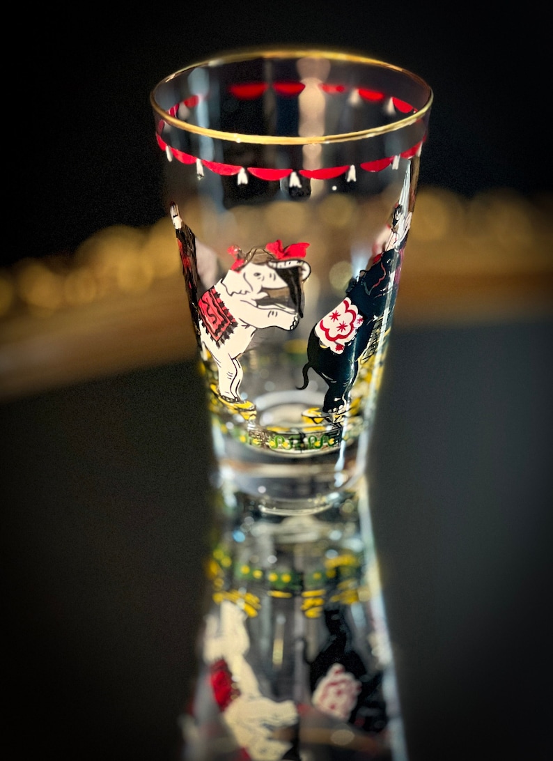 May include: A clear glass drinking glass with a gold rim and a circus-themed design. The design features two elephants, one white and one black, with red and yellow accents. The elephants are wearing colorful costumes and are standing on a green and yellow patterned background.