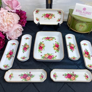 May include: A set of seven white porcelain trinket trays with gold trim and a floral design featuring red roses. The trays are in various sizes and shapes, including rectangular and oval. The trays are arranged on a black surface. The brand name "Royal Albert England" is visible on a small sign.