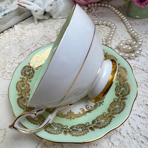 May include: A white teacup with a gold rim and handle rests on a light green saucer with gold filigree detailing. The teacup and saucer are part of a vintage tea set. The handle of the teacup has a heart shape.