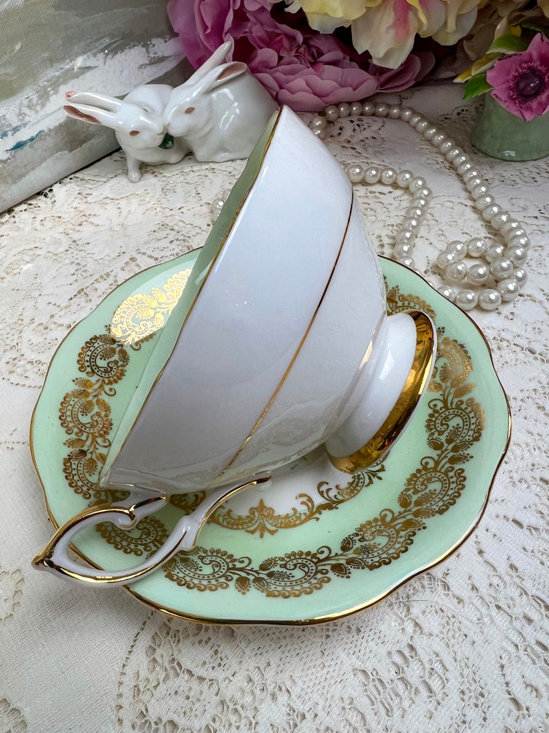 May include: A white teacup with a gold rim and handle rests on a light green saucer with gold filigree detailing. The teacup and saucer are part of a vintage tea set. The handle of the teacup has a heart shape.